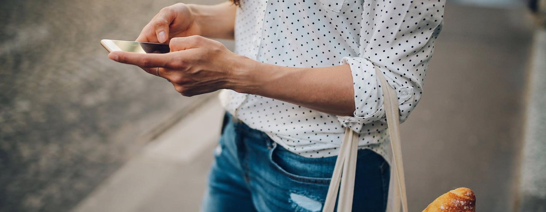 woman walks down the street and texts with a bag of groceries on her arm